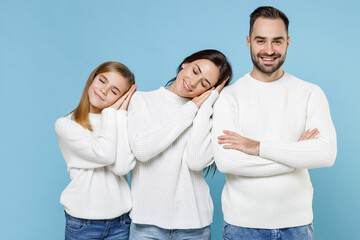 Smiling young parents mom dad with child kid daughter teen girl in sweaters sleep with folded hands under cheek isolated on blue background studio portrait. Family day parenthood childhood concept.