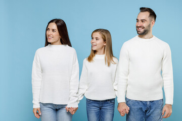 Obraz premium Funny young parents mom dad with child kid daughter teen girl in basic white sweaters holding hands looking aside isolated on blue background studio portrait. Family day parenthood childhood concept.