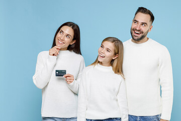 Pensive young parents mom dad with child kid daughter teen girl in white sweaters hold credit bank card looking aside up isolated on blue background studio portrait. Family day parenthood concept.