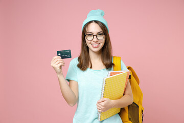 Smiling young woman student in casual blue t-shirt hat glasses backpack hold notebooks credit bank card isolated on pastel pink color background. Education in high school university college concept.