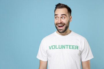Excited young bearded man in white volunteer t-shirt standing keeping mouth open looking aside isolated on blue background studio portrait. Voluntary free work assistance help charity grace concept.