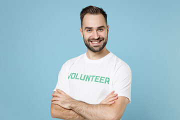 Smiling funny young bearded man 20s wearing white volunteer t-shirt holding hands crossed isolated...
