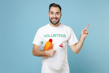 Smiling young man in volunteer t-shirt hold plastic bottles trash pointing index finger aside up isolated on blue background studio. Voluntary free assistance help trash sorting recycling concept.