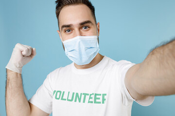 Close up of man in volunteer t-shirt mask to safe from coronavirus covid-19 doing selfie on mobile phone winner gesture isolated on blue background. Voluntary work assistance aid help support concept.