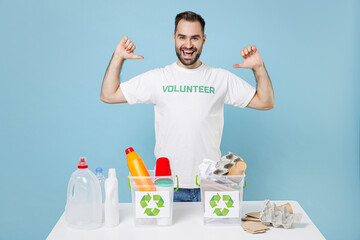 Funny young man in volunteer t-shirt stand near recycling stations sorting plastic paper trash point thumbs on himself isolated on blue background. Voluntary free work assistance help grace concept.