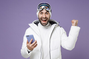Happy skier man in white windbreaker jacket ski goggles mask using mobile phone doing winner...