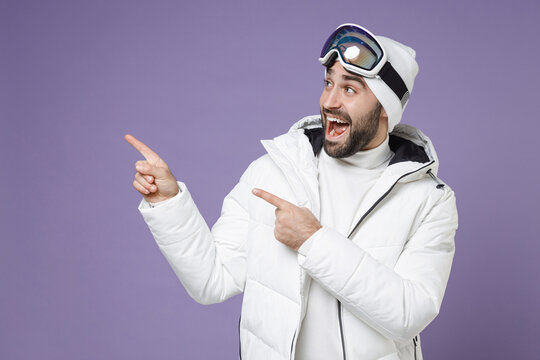 Excited skier man in white windbreaker jacket ski goggles mask pointing index fingers aside up spend extreme weekend winter in mountains isolated on purple background. People lifestyle hobby concept.