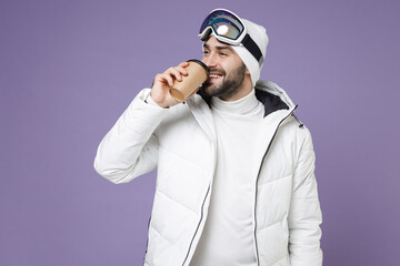 Funny skier man in white windbreaker jacket ski goggles mask hold paper cup coffee tea drinking spend extreme weekend winter in mountains isolated on purple background. People lifestyle hobby concept.