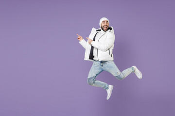 Full length of excited man in warm white padded windbreaker jacket hat jumping pointing index fingers aside isolated on purple background studio portrait. People lifestyle cold winter season concept. © ViDi Studio