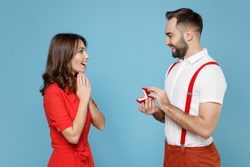 Side view of shocked young couple friends man woman in white red clothes going to propose marriage hold box with wedding ring isolated on blue background in studio. Valentine's Day holiday concept.