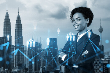 Smiling black woman HR director at international company is thinking about recruitment of highly qualified specialists. Women in business concept. Social media hologram icons over Kuala Lumpur.
