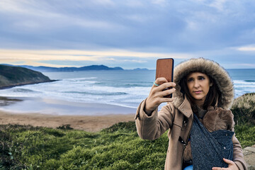 woman taking a selfie with the sea and the beach in the background in winter carrying her baby....