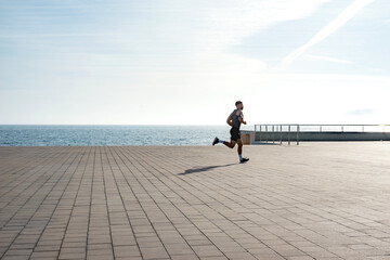 Fit young man running along seafront. Male runner jogging early in the morning. Fitness training outdoors. Workout during lockdown outside the gym.