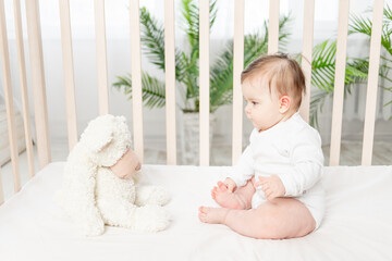 baby six months sitting in a crib in a white bodysuit with toys Teddy bears