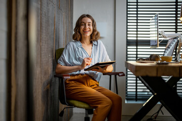 Portrait of a young woman as a digital artist, drawing on a digital tablet at the cozy workplace at...