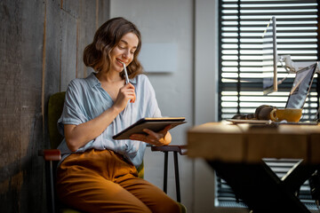 Young woman dressed casually having some creative work, drawing on a digital tablet, sitting at the cozy and stylish home office