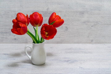 Bouquet of beautiful fresh red tulips in small white jug on light wooden background. Valentine's, women's or mother's day concept. Greeting card for spring holiday. Still life, selective focus