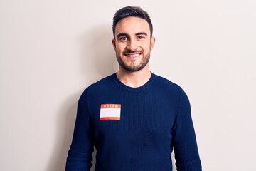 Young handsome man with beard wearing identification sticker over isolated white background looking positive and happy standing and smiling with a confident smile showing teeth