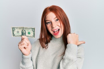 Young red head girl holding 1 dollar banknote pointing thumb up to the side smiling happy with open mouth