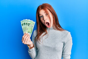 Young red head girl holding 10000 south korean won banknotes scared and amazed with open mouth for...
