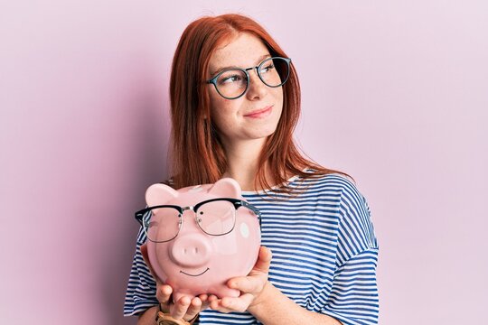 Young red head girl holding piggy bank with glasses smiling looking to the side and staring away thinking.