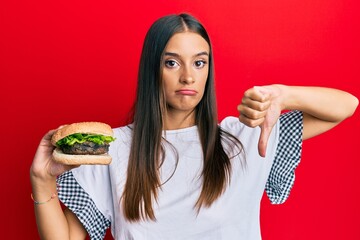 Young hispanic woman eating hamburger with angry face, negative sign showing dislike with thumbs down, rejection concept