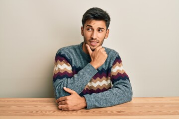 Fototapeta premium Young handsome man wearing casual sweater sitting on the table with hand on chin thinking about question, pensive expression. smiling with thoughtful face. doubt concept.