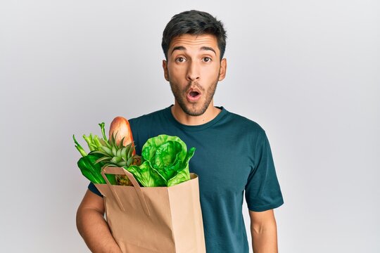 Young handsome man holding paper bag with bread and groceries scared and amazed with open mouth for surprise, disbelief face