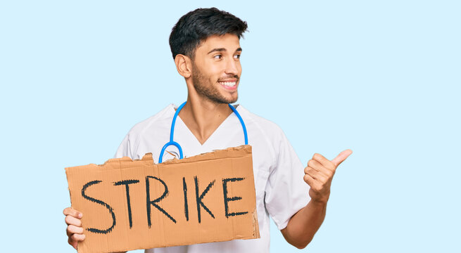 Young handsome man wearing doctor uniform holding strike banner cardboard pointing thumb up to the side smiling happy with open mouth