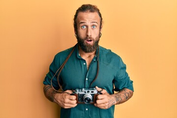 Handsome man with beard and long hair holding vintage camera afraid and shocked with surprise and amazed expression, fear and excited face.