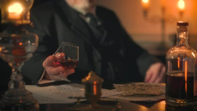 A Victorian era gentleman lit by candle and oil lamp light relaxing at a desk drinking liquor from a small brandy snifter check the time on his pocket watch.