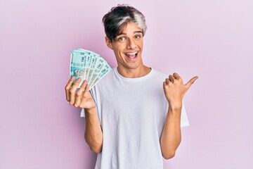 Young hispanic man holding 50 indian rupee banknotes pointing thumb up to the side smiling happy with open mouth
