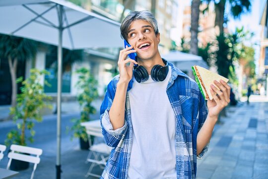 Young hispanic man smiling happy talking on the smartphone and eating sandwich at the city.