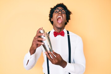 Handsome african american bartender man with afro hair preparing cocktail mixing drink with shaker angry and mad screaming frustrated and furious, shouting with anger looking up.