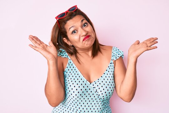 Middle age brunette hispanic woman wearing summer dress and sunglasses clueless and confused with open arms, no idea and doubtful face.