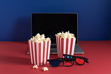 Laptop, popcorn in a red striped buckets and 3d glasses on red and blue background.