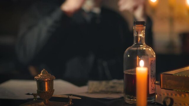 A Victorian era businessman lit by candle and oil lamp light relaxing at his desk drinking liquor from a small brandy snifter.
