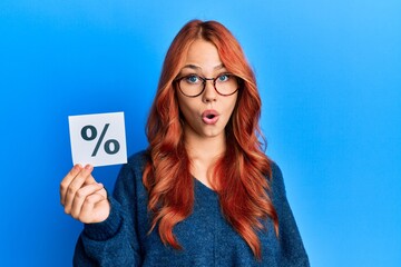 Young beautiful redhead woman holding percentage symbol scared and amazed with open mouth for...
