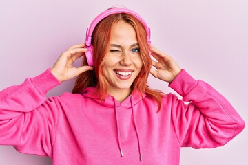 Young redhead woman listening to music using headphones winking looking at the camera with sexy...