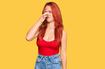 Young redhead woman wearing casual clothes smelling something stinky and disgusting, intolerable...