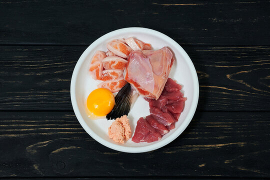 Natural raw cat food in bowl on black wooden background. Salmon fish, meat, egg and food supplements.