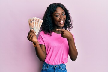 Beautiful african young woman holding 10 united kingdom pounds banknotes smiling happy pointing with hand and finger