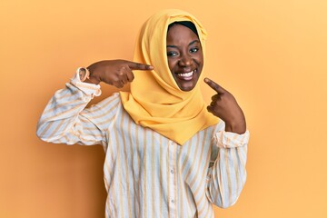 Beautiful african young woman wearing traditional islamic hijab scarf smiling cheerful showing and pointing with fingers teeth and mouth. dental health concept.