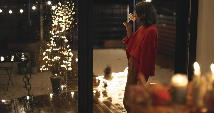 Woman standing with a drink by the window in the backyard of her house, celebrating winter holidays alone at night