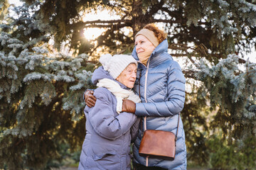 Concept of happy family, old age, emotions, senior care in retirement age. Active senior grandmother and adult daughter hugging outside in winter against background of Christmas tree in forest