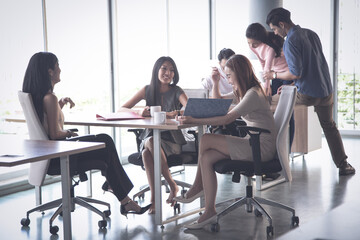 Group of young Asian business people working and communicating together while sitting at the office desk together with colleagues sitting in the background.