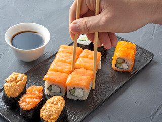 a variety of Japanese rolls and sushi on a textured black plate. Side view. Bamboo chopsticks holding a single roll