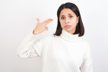 Unhappy Young brunette woman wearing white knitted sweater against white background makes suicide gesture and imitates gun with hand, curves lips, keeps two fingers on temple