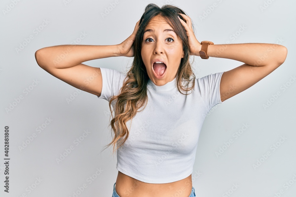 Wall mural young brunette woman wearing casual white t shirt crazy and scared with hands on head, afraid and su