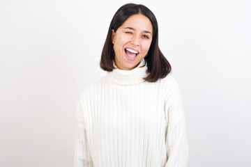 Young brunette woman wearing white knitted sweater against white background winking looking at the camera with sexy expression, cheerful and happy face.